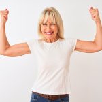 Middle age woman wearing casual t-shirt standing over isolated white background showing arms muscles smiling proud. Fitness concept.